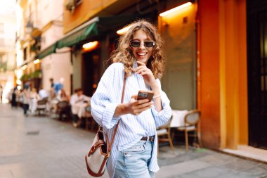Telefonu olan neşeli bir kadın güneşli bir günde şehir sokaklarında geziniyor. Güzel bir turist güneşli bir günün ve telefonuyla blog yazmanın tadını çıkarır. Turizm, tatil ve hafta sonları için konseptler.