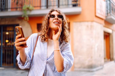 Telefonu olan neşeli bir kadın güneşli bir günde şehir sokaklarında geziniyor. Güzel bir turist güneşli bir günün ve telefonuyla blog yazmanın tadını çıkarır. Turizm, tatil ve hafta sonları için konseptler.