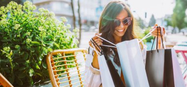 Alışveriş poşetleriyle gülümseyen bir kadın bir kafein terasında oturur. Güzel bir kadın alışverişten sonra güneşli bir günün tadını çıkarır. İndirim, alışveriş ve tüketim kavramı.