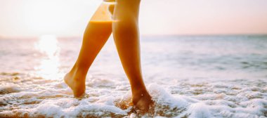 Yakın plan, genç bir kadın yaz sahilinde deniz suyu ve kum dalgası boyunca yürüyor..