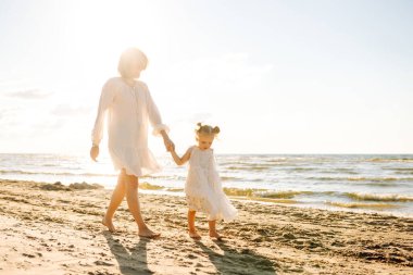 Happy mother and daughter are having fun on beach by water on sunny day. Beautiful woman and daughter are enjoying time together, spending time outdoors. Concepts of childhood and motherhood.