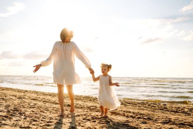 Happy mother and daughter are having fun on beach by water on sunny day. Beautiful woman and daughter are enjoying time together, spending time outdoors. Concepts of childhood and motherhood.