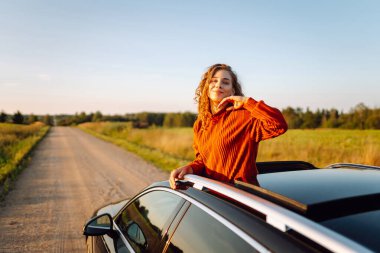 Araba penceresinden ya da çatısından sarkan bir kadın gezgin gün batımının özgürlüğünü hisseder. Yolculukta olan bir kadın güneşli bir günün tadını çıkarır. Macera konsepti, kırsal alan. Yaşam biçimi.