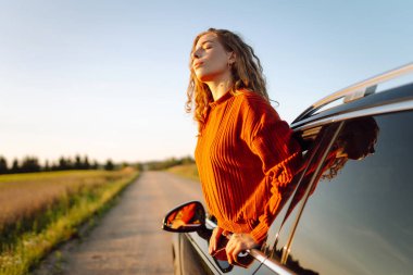 Araba penceresinden ya da çatısından sarkan bir kadın gezgin gün batımının özgürlüğünü hisseder. Yolculukta olan bir kadın güneşli bir günün tadını çıkarır. Macera konsepti, kırsal alan. Yaşam biçimi.