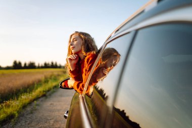 Araba penceresinden ya da çatısından sarkan bir kadın gezgin gün batımının özgürlüğünü hisseder. Yolculukta olan bir kadın güneşli bir günün tadını çıkarır. Macera konsepti, kırsal alan. Yaşam biçimi.