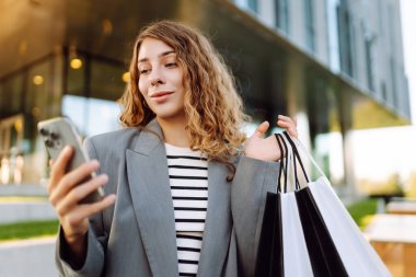 Güneşli bir günde modern bir binanın önünde elinde telefon ve alışveriş torbalarıyla duran genç bir kadının portresi. Şık bir kadın, internetten alışveriş yaptıktan sonra elinde telefon tutup açık hava alışverişi yapıyor. Tüketim kavramı.