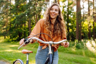 Genç bir kadın güneşli bir park yolu boyunca bisikletini sürüyor. Mutlu bir kadın ağaçların arasında bisiklet sürmekten hoşlanır. Doğa kavramı, hafta sonları ve rahatlama.