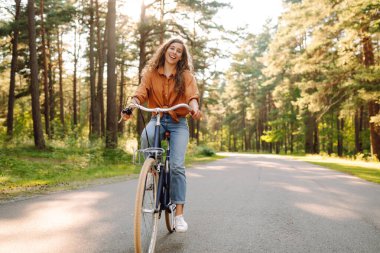 Genç bir kadın güneşli bir park yolu boyunca bisikletini sürüyor. Mutlu bir kadın ağaçların arasında bisiklet sürmekten hoşlanır. Doğa kavramı, hafta sonları ve rahatlama.