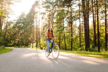 Genç bir kadın güneşli bir park yolu boyunca bisikletini sürüyor. Mutlu bir kadın ağaçların arasında bisiklet sürmekten hoşlanır. Doğa kavramı, hafta sonları ve rahatlama.
