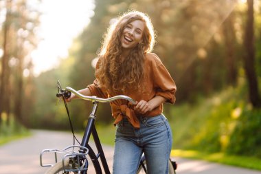 Genç bir kadın güneşli bir park yolu boyunca bisikletini sürüyor. Mutlu bir kadın ağaçların arasında bisiklet sürmekten hoşlanır. Doğa kavramı, hafta sonları ve rahatlama.