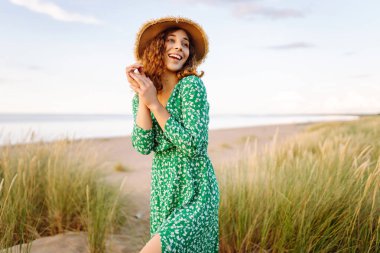 Hasır şapkalı mutlu bir kadın kumsalda günbatımının tadını çıkarıyor. Elbise giymiş genç bir kadın sahilde geziniyor ve denize bakıyor. Rahatlama, hafta sonları ve özgürlük kavramları.