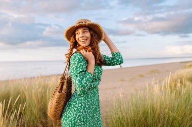 Hasır şapkalı mutlu bir kadın kumsalda günbatımının tadını çıkarıyor. Elbise giymiş genç bir kadın sahilde geziniyor ve denize bakıyor. Rahatlama, hafta sonları ve özgürlük kavramları.