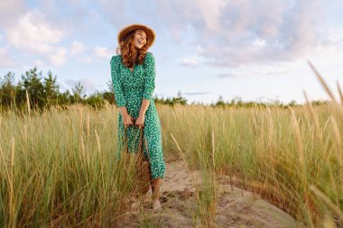 Hasır şapkalı mutlu bir kadın kumsalda günbatımının tadını çıkarıyor. Elbise giymiş genç bir kadın sahilde geziniyor ve denize bakıyor. Rahatlama, hafta sonları ve özgürlük kavramları.