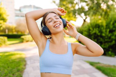 Kablosuz kulaklık takan genç sporcu güneşli parkta antrenmandan önce ısınıyor. Spor giyim sektöründe güzel bir kadın müzik dinler ve açık havayı sever. Konsept: eğlence, spor. Etkin yaşam biçimi.