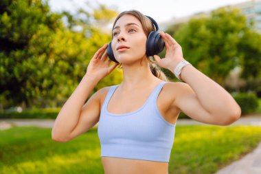 Kablosuz kulaklık takan genç sporcu güneşli parkta antrenmandan önce ısınıyor. Spor giyim sektöründe güzel bir kadın müzik dinler ve açık havayı sever. Konsept: eğlence, spor. Etkin yaşam biçimi.