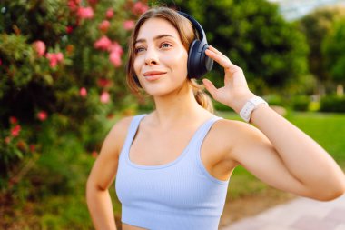 Kablosuz kulaklık takan genç sporcu güneşli parkta antrenmandan önce ısınıyor. Spor giyim sektöründe güzel bir kadın müzik dinler ve açık havayı sever. Konsept: eğlence, spor. Etkin yaşam biçimi.