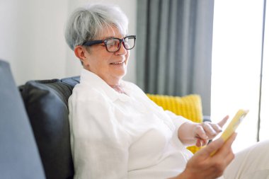 Daha yaşlı, gri saçlı bir kadın rahat bir odada bir koltukta oturuyor. Gülümseyen bir emekli, güneşli bir günde evde akıllı telefon kullanır. Mutlu emeklilik, teknoloji ve konfor kavramı.