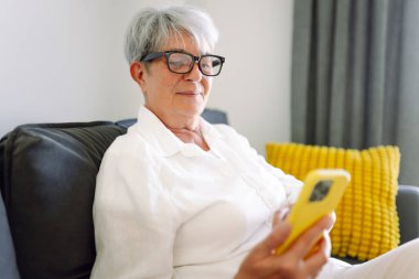 Daha yaşlı, gri saçlı bir kadın rahat bir odada bir koltukta oturuyor. Gülümseyen bir emekli, güneşli bir günde evde akıllı telefon kullanır. Mutlu emeklilik, teknoloji ve konfor kavramı.