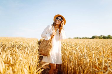 Beyaz elbiseli ve hasır şapkalı bir kadın güneşli bir günde altın buğday tarlasında geziniyor. Güzel kadın gün batımında özgür hissederek doğanın tadını çıkarıyor. Kavram: güzellik, tarz ve kırsal.