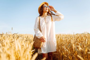 Beyaz elbiseli ve hasır şapkalı bir kadın güneşli bir günde altın buğday tarlasında geziniyor. Güzel kadın gün batımında özgür hissederek doğanın tadını çıkarıyor. Kavram: güzellik, tarz ve kırsal.