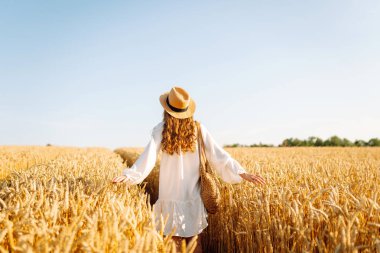 Beyaz elbiseli ve hasır şapkalı bir kadın güneşli bir günde altın buğday tarlasında geziniyor. Güzel kadın gün batımında özgür hissederek doğanın tadını çıkarıyor. Kavram: güzellik, tarz ve kırsal.