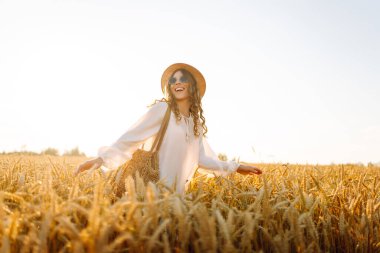 Beyaz elbiseli ve hasır şapkalı bir kadın güneşli bir günde altın buğday tarlasında geziniyor. Güzel kadın gün batımında özgür hissederek doğanın tadını çıkarıyor. Kavram: güzellik, tarz ve kırsal.