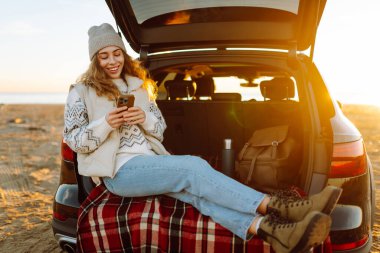 Mutlu kadın telefonu güneşli sahilde bir arabanın bagajında oturuyor. Yolculuk blogunda bir kadın ve deniz kıyısında gün doğumunun tadını çıkarıyor. Ulaşım, blog ve boş zaman kavramları.