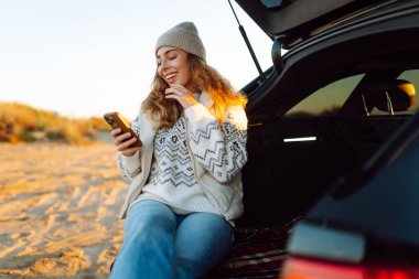 Mutlu kadın telefonu güneşli sahilde bir arabanın bagajında oturuyor. Yolculuk blogunda bir kadın ve deniz kıyısında gün doğumunun tadını çıkarıyor. Ulaşım, blog ve boş zaman kavramları.