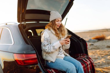 Mutlu kadın telefonu güneşli sahilde bir arabanın bagajında oturuyor. Yolculuk blogunda bir kadın ve deniz kıyısında gün doğumunun tadını çıkarıyor. Ulaşım, blog ve boş zaman kavramları.