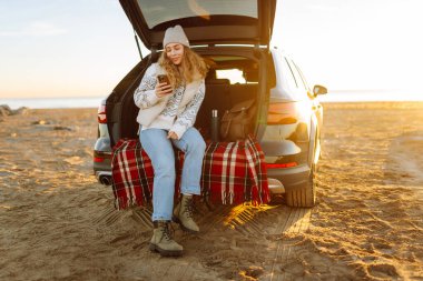 Mutlu kadın telefonu güneşli sahilde bir arabanın bagajında oturuyor. Yolculuk blogunda bir kadın ve deniz kıyısında gün doğumunun tadını çıkarıyor. Ulaşım, blog ve boş zaman kavramları.