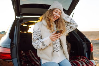 Mutlu kadın telefonu güneşli sahilde bir arabanın bagajında oturuyor. Yolculuk blogunda bir kadın ve deniz kıyısında gün doğumunun tadını çıkarıyor. Ulaşım, blog ve boş zaman kavramları.
