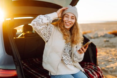 Mutlu kadın telefonu güneşli sahilde bir arabanın bagajında oturuyor. Yolculuk blogunda bir kadın ve deniz kıyısında gün doğumunun tadını çıkarıyor. Ulaşım, blog ve boş zaman kavramları.
