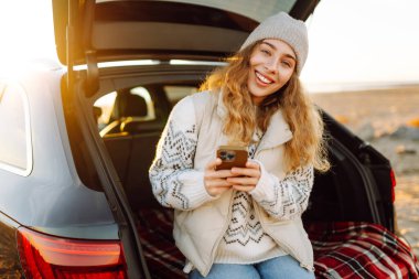 Mutlu kadın telefonu güneşli sahilde bir arabanın bagajında oturuyor. Yolculuk blogunda bir kadın ve deniz kıyısında gün doğumunun tadını çıkarıyor. Ulaşım, blog ve boş zaman kavramları.