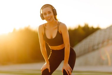 Atletik kadın koşudan ya da antrenmandan önce ısınıyor, güneş altında kablosuz kulaklık takıyor. Spor giyim sektöründeki kadınlar güneşli bir günün tadını çıkarıyorlar, müzik dinliyor ve egzersiz yapıyorlar. Spor anlayışı ve esneme.