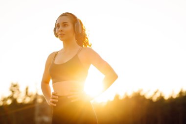 Atletik kadın koşudan ya da antrenmandan önce ısınıyor, güneş altında kablosuz kulaklık takıyor. Spor giyim sektöründeki kadınlar güneşli bir günün tadını çıkarıyorlar, müzik dinliyor ve egzersiz yapıyorlar. Spor anlayışı ve esneme.