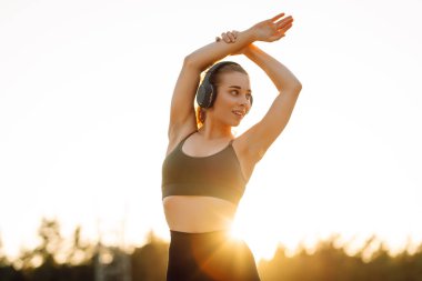 Atletik kadın koşudan ya da antrenmandan önce ısınıyor, güneş altında kablosuz kulaklık takıyor. Spor giyim sektöründeki kadınlar güneşli bir günün tadını çıkarıyorlar, müzik dinliyor ve egzersiz yapıyorlar. Spor anlayışı ve esneme.