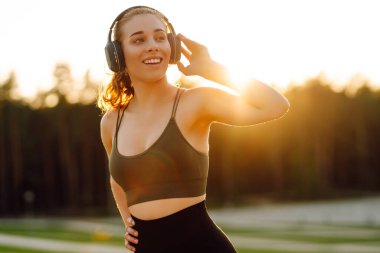 Atletik kadın koşudan ya da antrenmandan önce ısınıyor, güneş altında kablosuz kulaklık takıyor. Spor giyim sektöründeki kadınlar güneşli bir günün tadını çıkarıyorlar, müzik dinliyor ve egzersiz yapıyorlar. Spor anlayışı ve esneme.