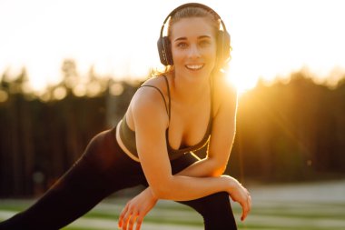 Atletik kadın koşudan ya da antrenmandan önce ısınıyor, güneş altında kablosuz kulaklık takıyor. Spor giyim sektöründeki kadınlar güneşli bir günün tadını çıkarıyorlar, müzik dinliyor ve egzersiz yapıyorlar. Spor anlayışı ve esneme.