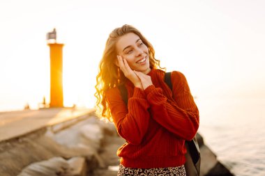 Sırt çantalı genç bir kadın deniz fenerine doğru yürüyüp günbatımının tadını çıkarıyor. Gezgin açık havanın özgürlüğünü hisseder. Yürüme, hafta sonları ve özgürlük kavramları.