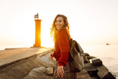 Sırt çantalı genç bir kadın deniz fenerine doğru yürüyüp günbatımının tadını çıkarıyor. Gezgin açık havanın özgürlüğünü hisseder. Yürüme, hafta sonları ve özgürlük kavramları.