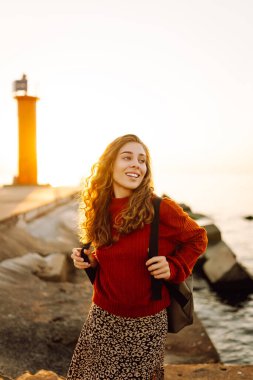 Sırt çantalı genç bir kadın deniz fenerine doğru yürüyüp günbatımının tadını çıkarıyor. Gezgin açık havanın özgürlüğünü hisseder. Yürüme, hafta sonları ve özgürlük kavramları.