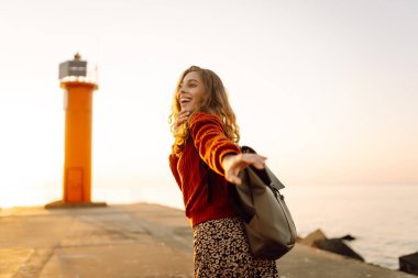 Sırt çantalı genç bir kadın deniz fenerine doğru yürüyüp günbatımının tadını çıkarıyor. Gezgin açık havanın özgürlüğünü hisseder. Yürüme, hafta sonları ve özgürlük kavramları.