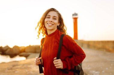 Sırt çantalı genç bir kadın deniz fenerine doğru yürüyüp günbatımının tadını çıkarıyor. Gezgin açık havanın özgürlüğünü hisseder. Yürüme, hafta sonları ve özgürlük kavramları.