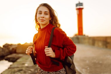 Sırt çantalı genç bir kadın deniz fenerine doğru yürüyüp günbatımının tadını çıkarıyor. Gezgin açık havanın özgürlüğünü hisseder. Yürüme, hafta sonları ve özgürlük kavramları.