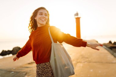 Sırt çantalı genç bir kadın deniz fenerine doğru yürüyüp günbatımının tadını çıkarıyor. Gezgin açık havanın özgürlüğünü hisseder. Yürüme, hafta sonları ve özgürlük kavramları.