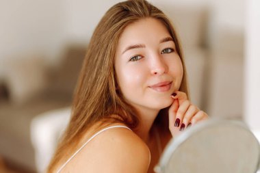 Tatlı kadın küçük bir aynanın önünde oturur, gün ışığında parmaklarıyla yüzüne dokunur. Güzel bir kadın rahat bir odada doğal güzelliğin ve gençliğin tadını çıkarır. Güzellik, tımar ve gençlik kavramları.