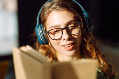 Kablosuz kulaklık takan, rahat bir odada kitap okuyan bir kadının kaybolması. Gözlüklü öğrenci kitap okumayı ve gün ışığında dinlenmeyi sever. Eğitimin kavramsal kavramları, rahatlama, teknoloji.