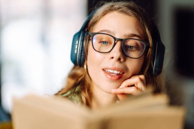 Kablosuz kulaklık takan, rahat bir odada kitap okuyan bir kadının kaybolması. Gözlüklü öğrenci kitap okumayı ve gün ışığında dinlenmeyi sever. Eğitimin kavramsal kavramları, rahatlama, teknoloji.