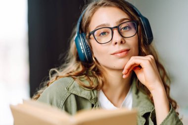 Kablosuz kulaklık takan, rahat bir odada kitap okuyan bir kadının kaybolması. Gözlüklü öğrenci kitap okumayı ve gün ışığında dinlenmeyi sever. Eğitimin kavramsal kavramları, rahatlama, teknoloji.