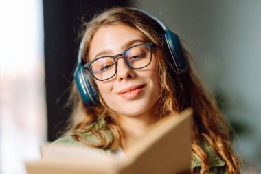 Kablosuz kulaklık takan, rahat bir odada kitap okuyan bir kadının kaybolması. Gözlüklü öğrenci kitap okumayı ve gün ışığında dinlenmeyi sever. Eğitimin kavramsal kavramları, rahatlama, teknoloji.